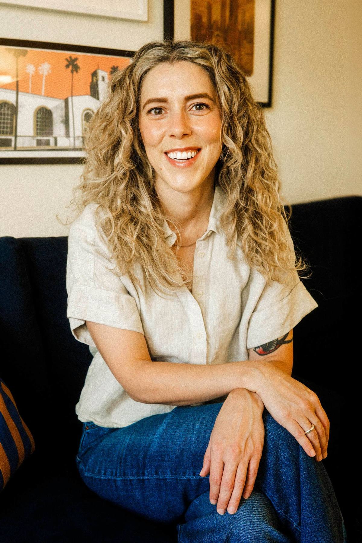 Nikole Sparks smiling, with long curly blonde hair, wearing a cream linen shirt and jeans, seated on a navy sofa